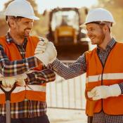 Two construction workers in orange vests and white hard hands shake hands while smiling