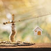 A crane hoisting up a model home sits on a stack of gold coins, indicating the construction loans needed to build housing.