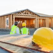 Two home builders stand facing under-construction home. A hard hat sits in the foreground. 