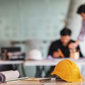 A student and teacher go over an assignment in the background. In the foreground, there is a table with some papers and a hard hat. 