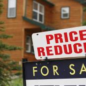 A "For Sale" sign with a "Price Reduced" sign added above it sit in front of an older home