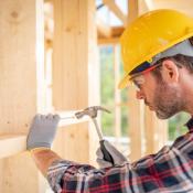 A man in a yellow hard hat hammers a nail into the wall of a home