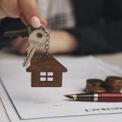 A real estate agent hands over a house key to a buyer. On the desk where they are seated, there is a contract with a pen, indicating it was recently signed.