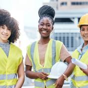 Three young women stand at a construction site wearing yellow vests 