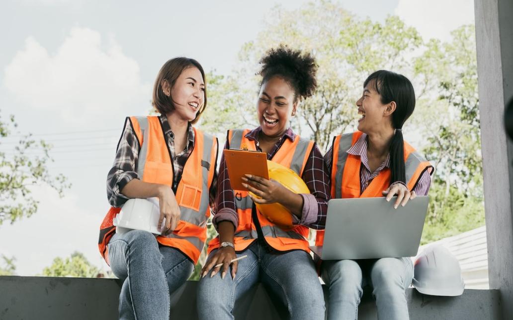 Three women in orange vests sitting, laughing together, hard hat sits to the right of the image