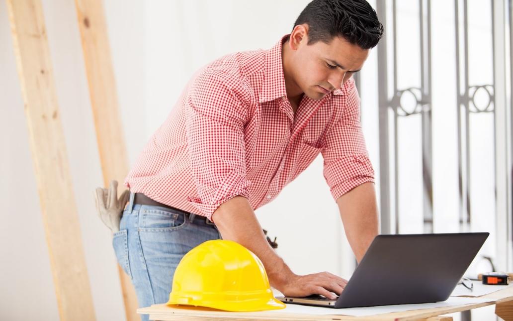 A young construction worker types on a laptop. A yellow hard hat sits off to the side.