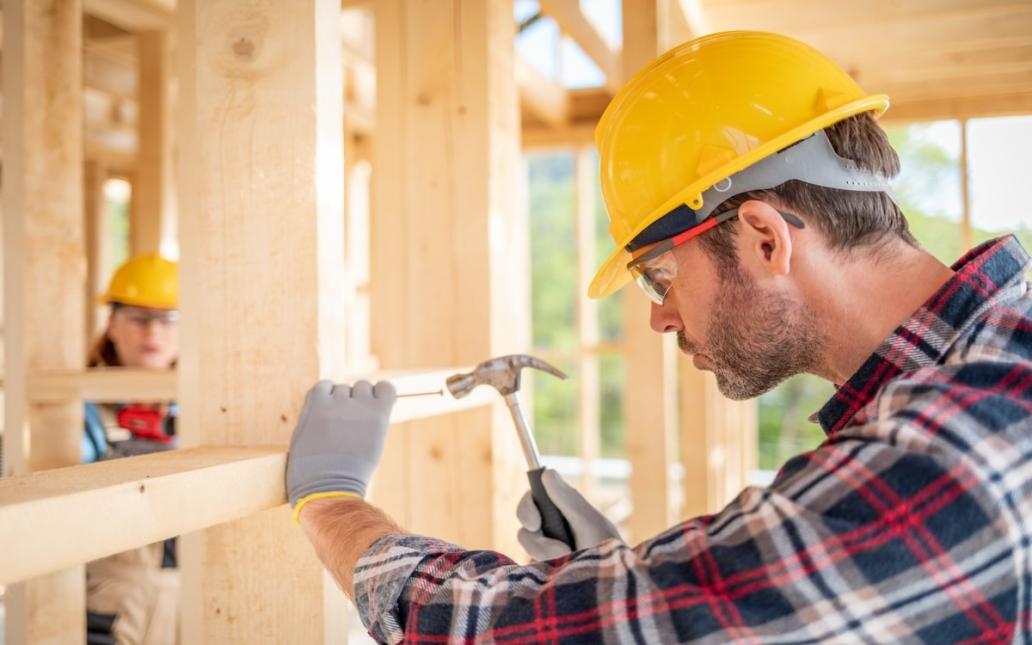 A man in a yellow hard hat hammers a nail into the wall of a home