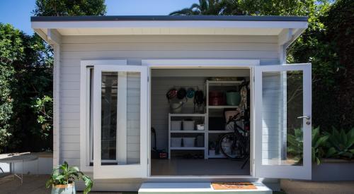 Accessory dwelling unit sits in a backyard with door open revealing interior shelving unit