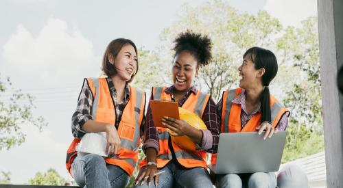 Three women in orange vests sitting, laughing together, hard hat sits to the right of the image