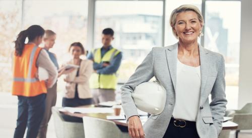 Woman in blazer smiles and holds hard hat, behind her are four construction workers conversing