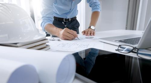 A man in business casual clothing works on construction plans. A hard hat, laptop and several rolls of paper sit around the plans on a table