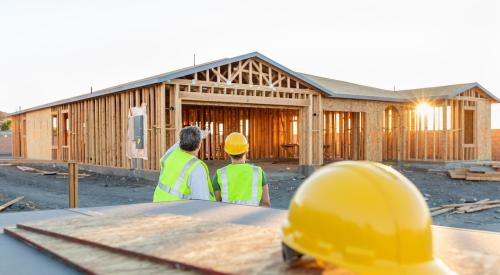 Two home builders stand facing under-construction home. A hard hat sits in the foreground. 