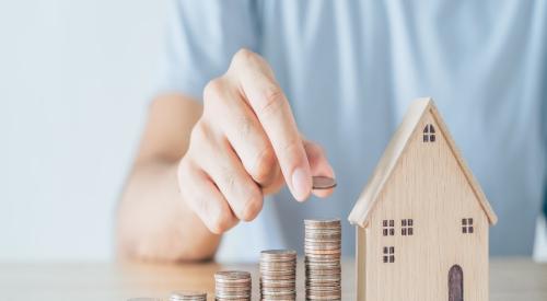 An individual sits at a table, stacking rows of coins in height ascending order. A wooden model home sits next to the coins, indicating home price growth. 