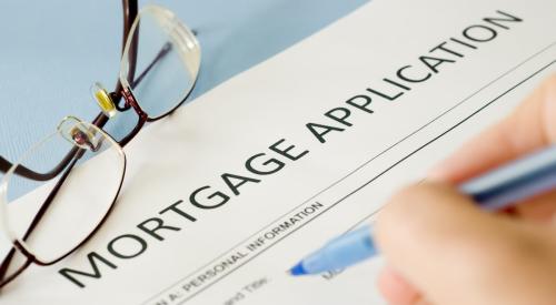 A homebuyer fills out a mortgage application with a blue pen. A pair of glasses sit off to the side of the application