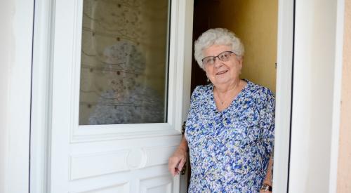 An elderly woman stands at the front door of her home