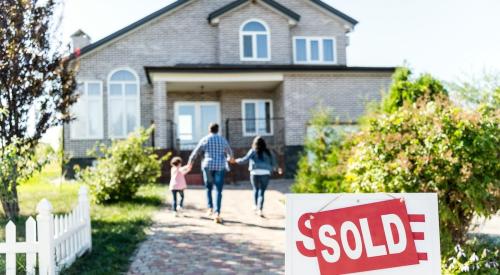 A family holds hands, running toward a single-family home with a "Sold" sign in the front yard