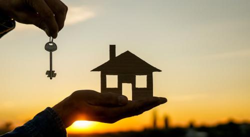 Person holding house cutout and keys against sunset sky